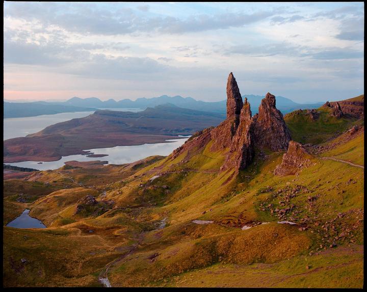 A dramatic landscape shows a large rock formation on a grassy hill overlooking a body of water and distant mountains under a cloudy sky.