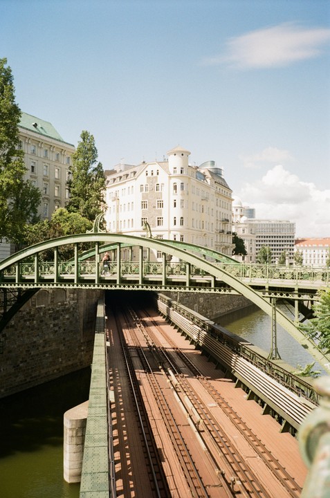 A green metal bridge with a pedestrian and a cyclist crosses over train tracks and a river, with buildings and trees in the background.