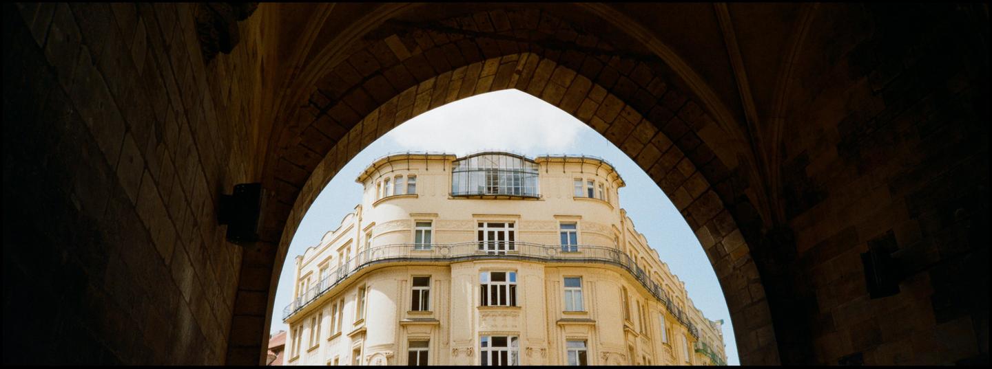 A large, light-colored building with many windows is framed by a dark stone archway.