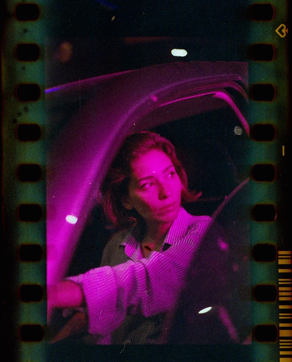 A woman sits in a car at night, illuminated by pink light.