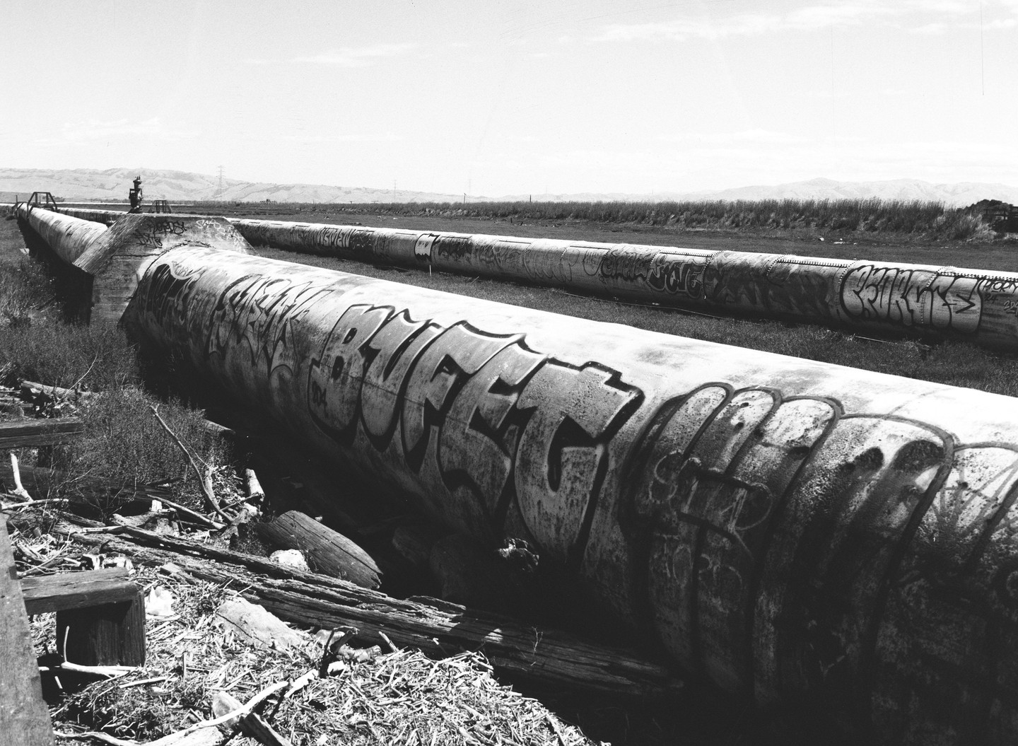 Two large pipes covered in graffiti run through a grassy field towards distant hills under a bright sky.