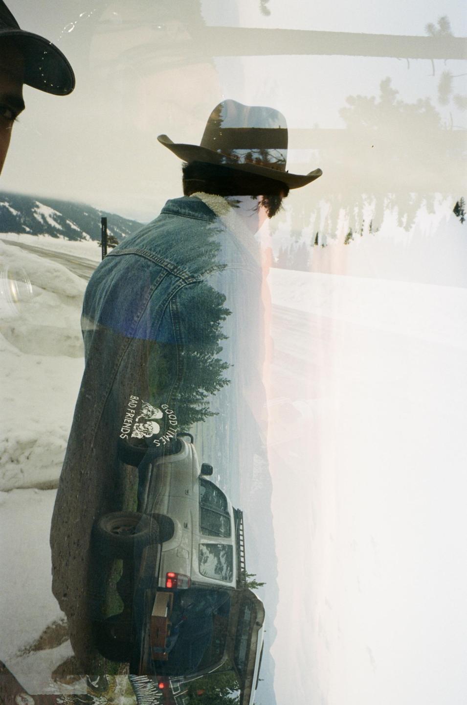 A man in a cowboy hat and denim jacket stands in a snowy landscape, with a double exposure of a car and trees superimposed on the image.