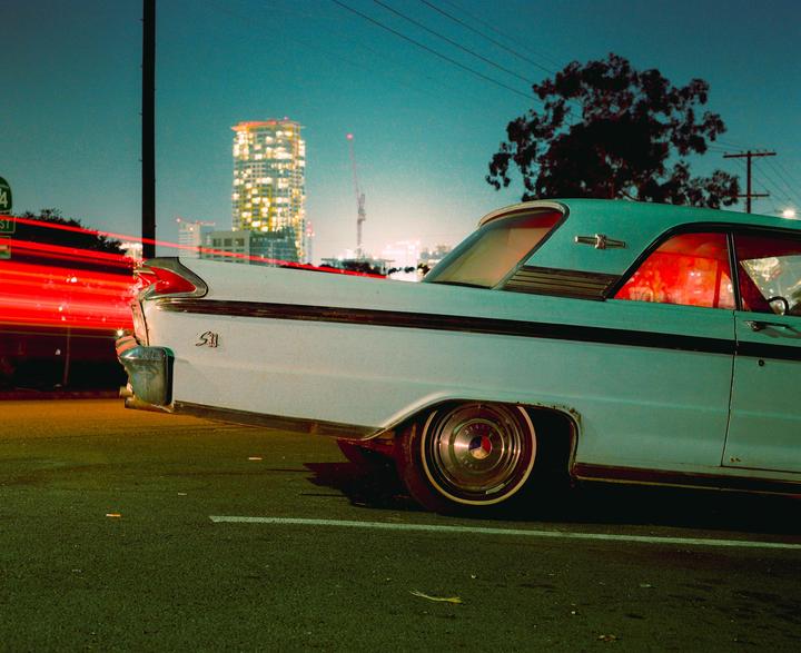 A light blue vintage car is parked on a street at night with a city skyline in the background and red light trails from passing vehicles.