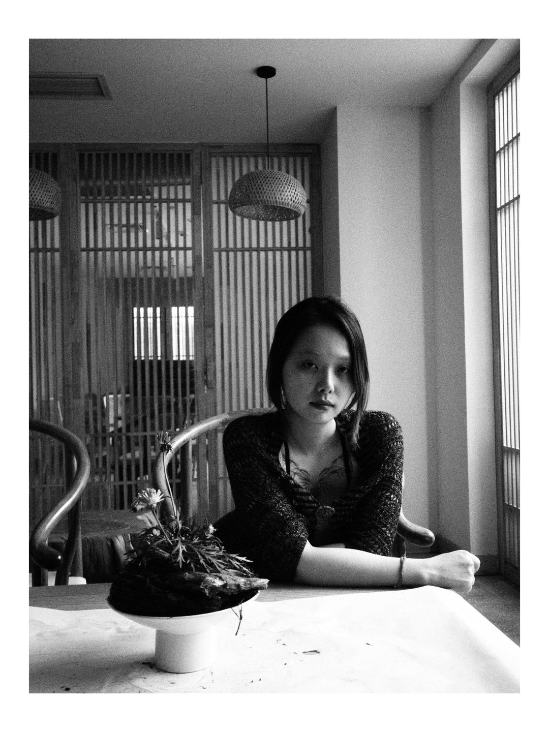A woman sits at a table with her arms crossed, looking directly at the viewer.
