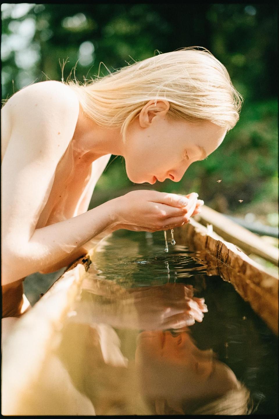 A woman with blonde hair is cupping her hands to drink water from a wooden trough, with her reflection visible in the water.