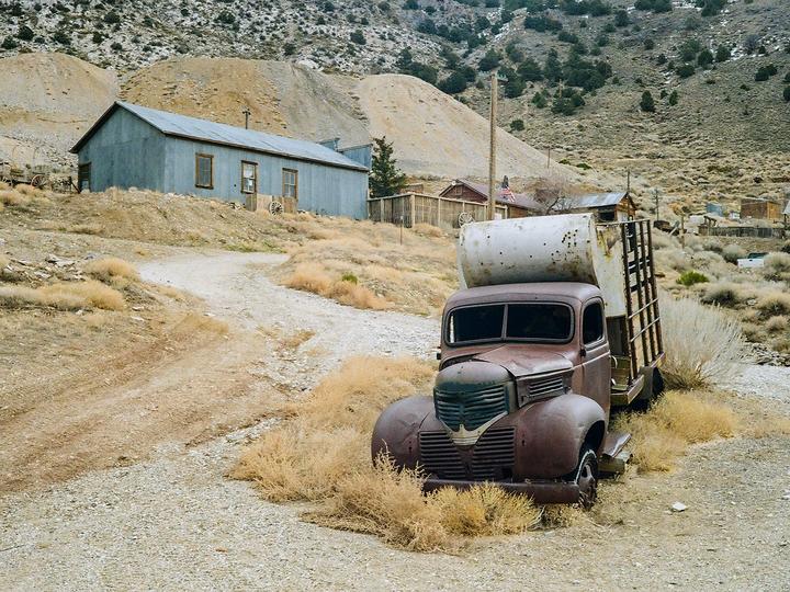 A rusty old truck with a tank in its bed sits on a dirt road in a desert landscape with a building and hills in the background.