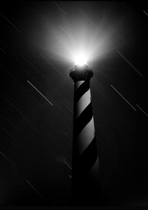 A black and white image shows a lighthouse with a bright beam of light against a night sky with star trails.
