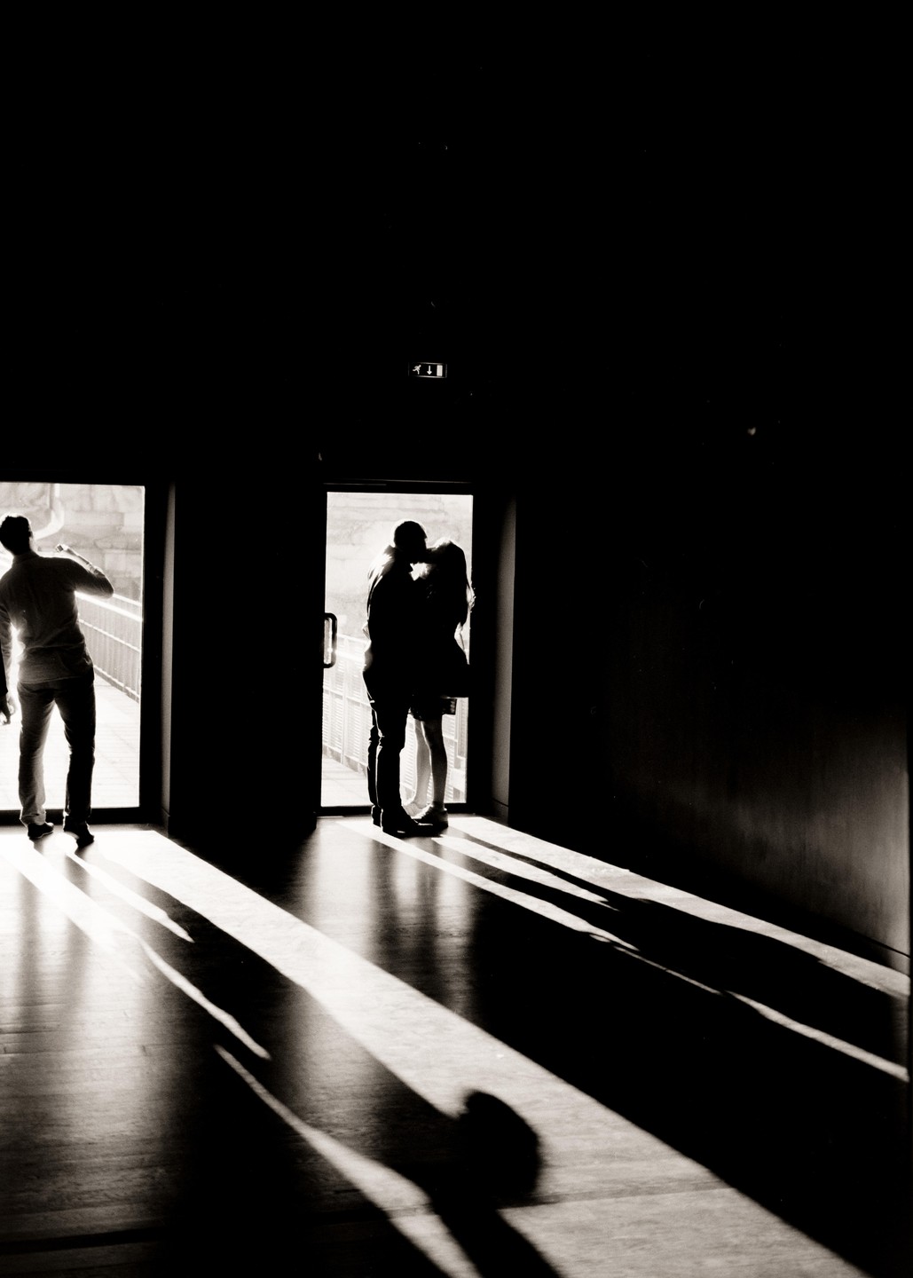 A man stands in a doorway while a couple kisses in another doorway, casting long shadows on the floor.