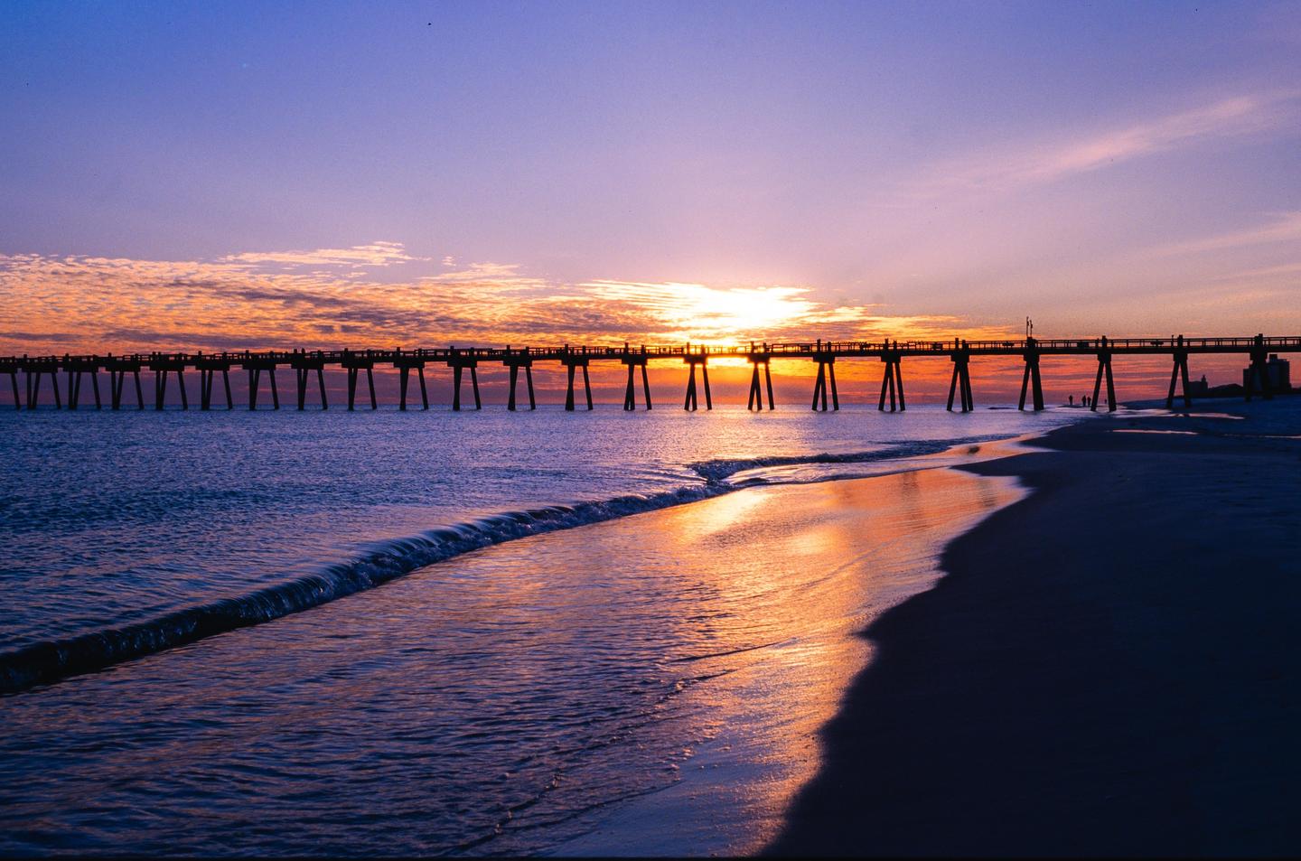 A long pier stretches across the water under a colorful sunset sky with waves breaking on the beach.