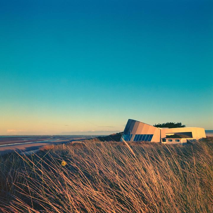 A modern building with a sloped roof stands on a grassy dune overlooking a beach at sunset.