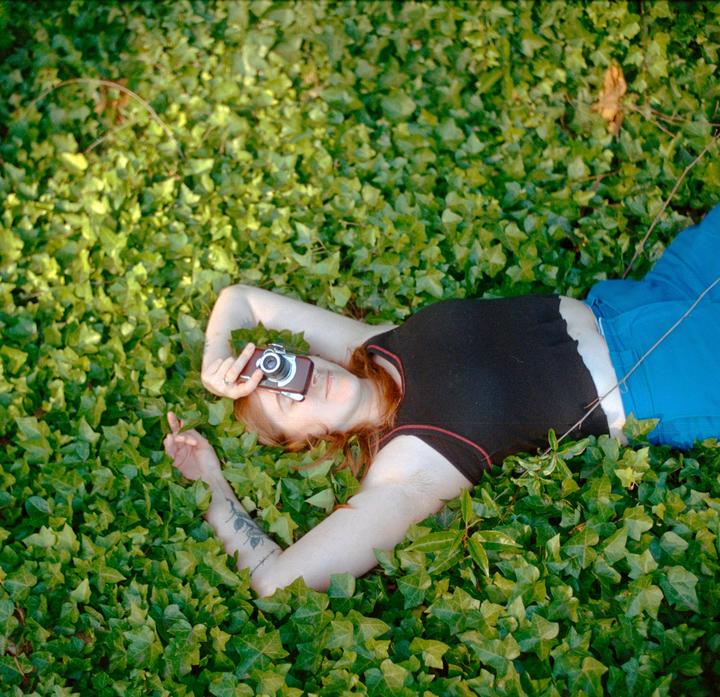 A woman with red hair lies on her back in a bed of ivy, holding a camera over her face.