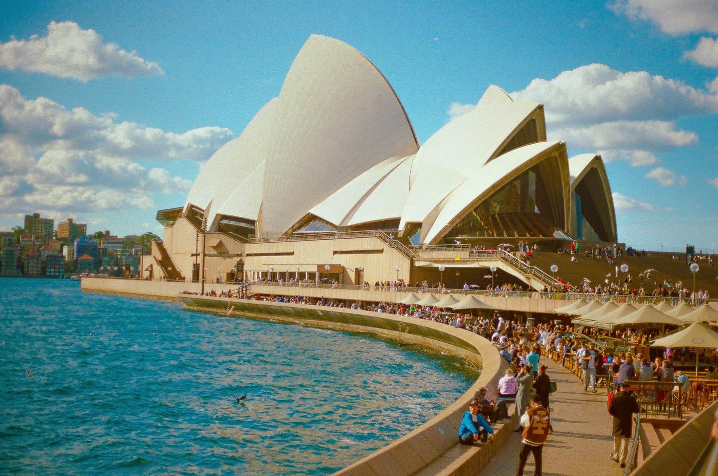 The Sydney Opera House is visible from the water, with many people gathered on the promenade and a city skyline in the distance.