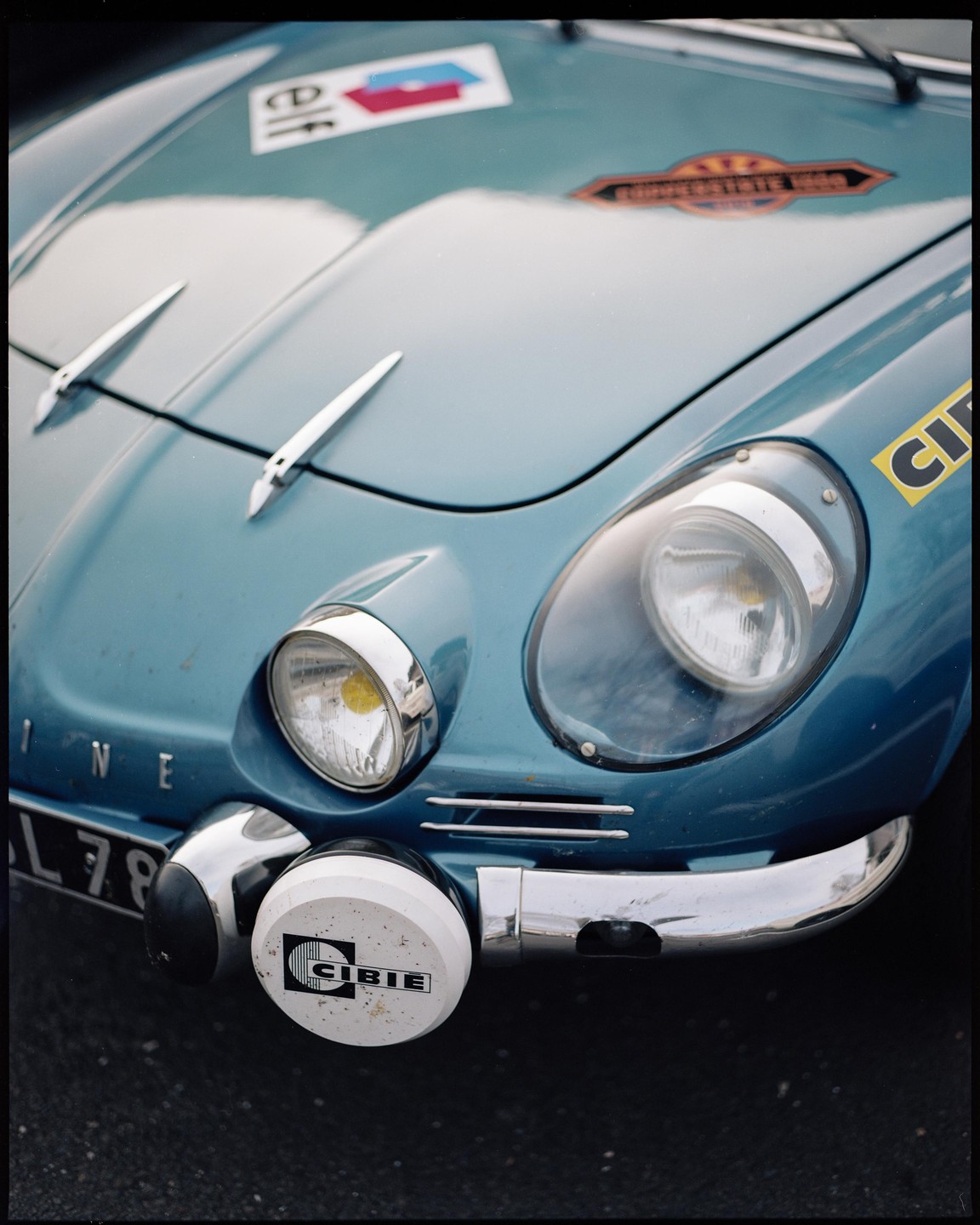 The front of a blue vintage sports car with rally stickers and auxiliary lights is shown.