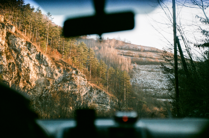 A view from inside a car shows a mountainous landscape with trees and patches of snow.