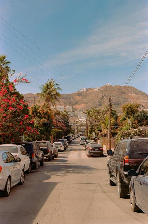 A street lined with parked cars leads towards a mountain with buildings on its slopes under a clear sky.
