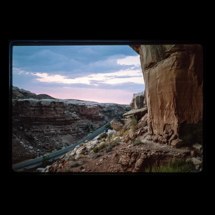 A road winds through a canyon with large rock formations under a cloudy sky at dusk.