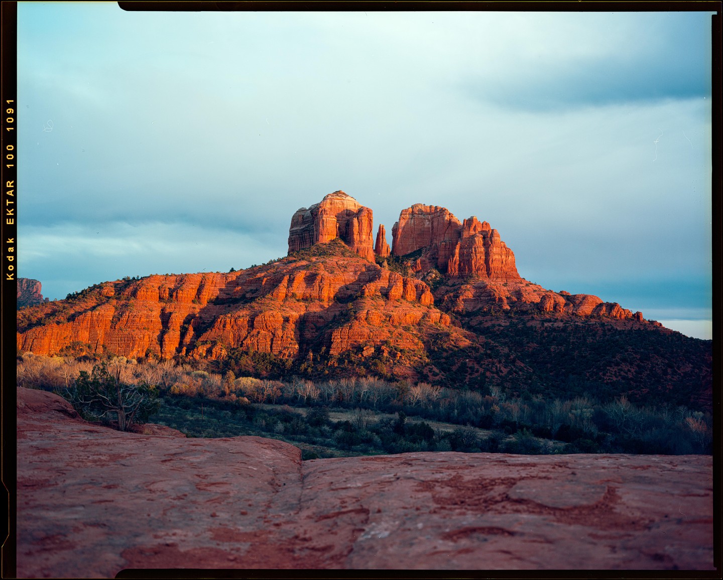A large red rock formation is illuminated by the setting sun under a blue sky with clouds.