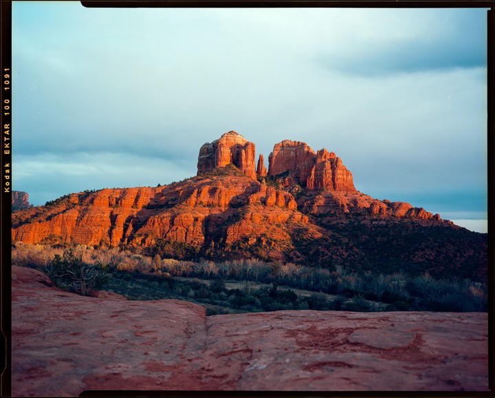 A large red rock formation is illuminated by the setting sun under a blue sky with clouds.