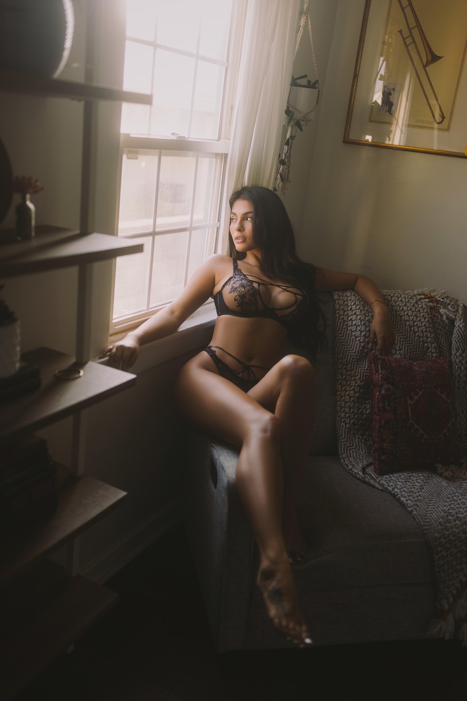A woman in lingerie sits on a chair next to a window, looking out.