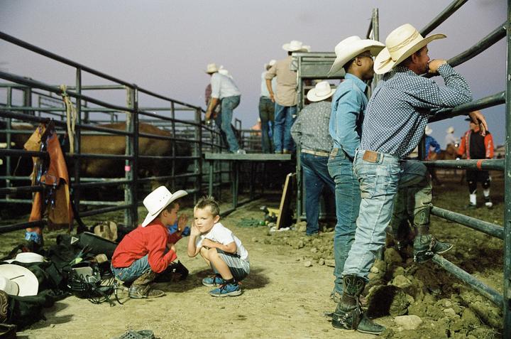 Two boys in cowboy hats squat on the ground near a metal fence while men in cowboy hats and jeans stand around a rodeo arena.