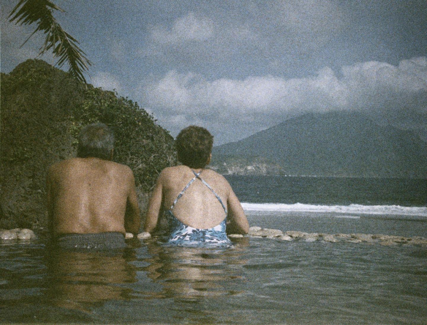 A couple sits in a pool of water looking out at the ocean and a distant mountain.