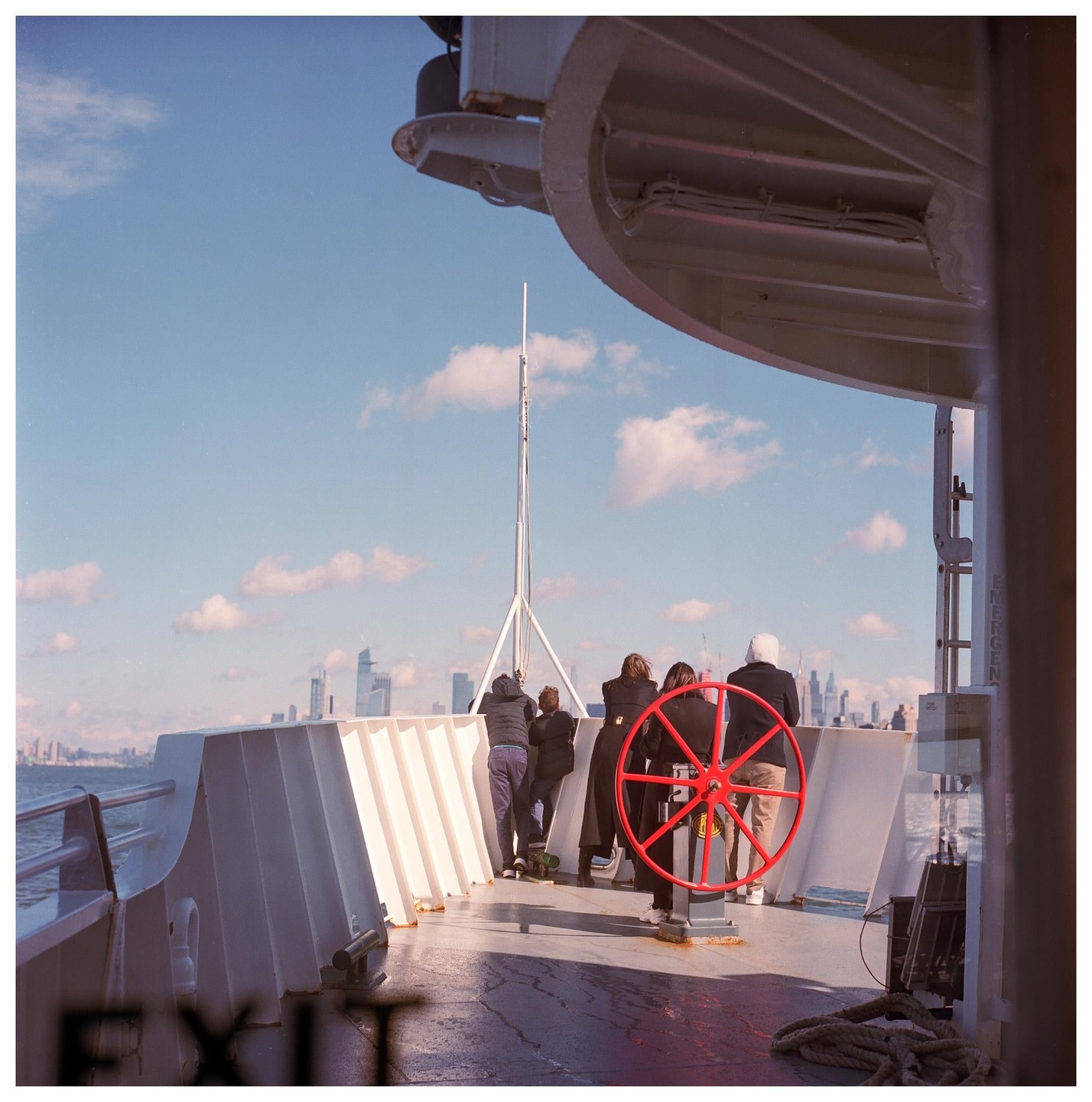 People stand on the deck of a ferry, looking out at a city skyline under a blue sky with clouds.