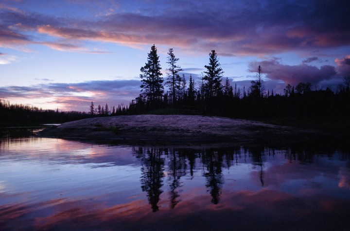 A body of water reflects the purple and pink hues of a sunset sky, with a dark tree-covered island in the middle ground.