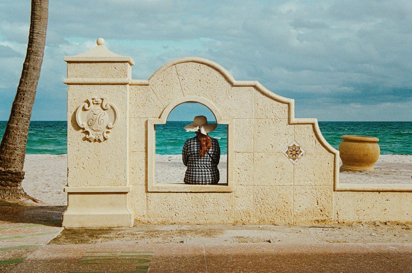 A woman with red hair and a hat sits in an arched opening of a stone wall, looking out at the ocean and beach.