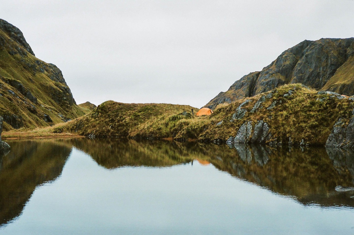 An orange tent is pitched on a grassy hill next to a calm body of water, with mountains rising in the background.