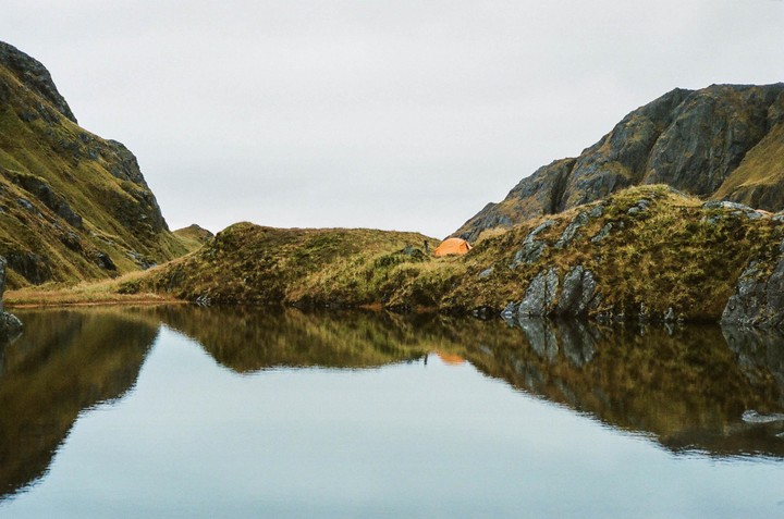 An orange tent is pitched on a grassy hill next to a calm body of water, with mountains rising in the background.