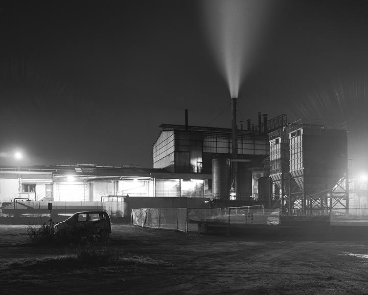 A plume of smoke rises from a factory smokestack at night, with a car parked in the foreground.