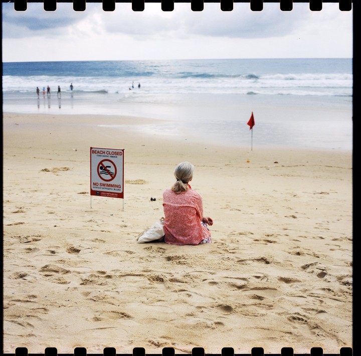 A woman sits on a sandy beach with a "beach closed" sign and a red flag nearby, looking out at the ocean.