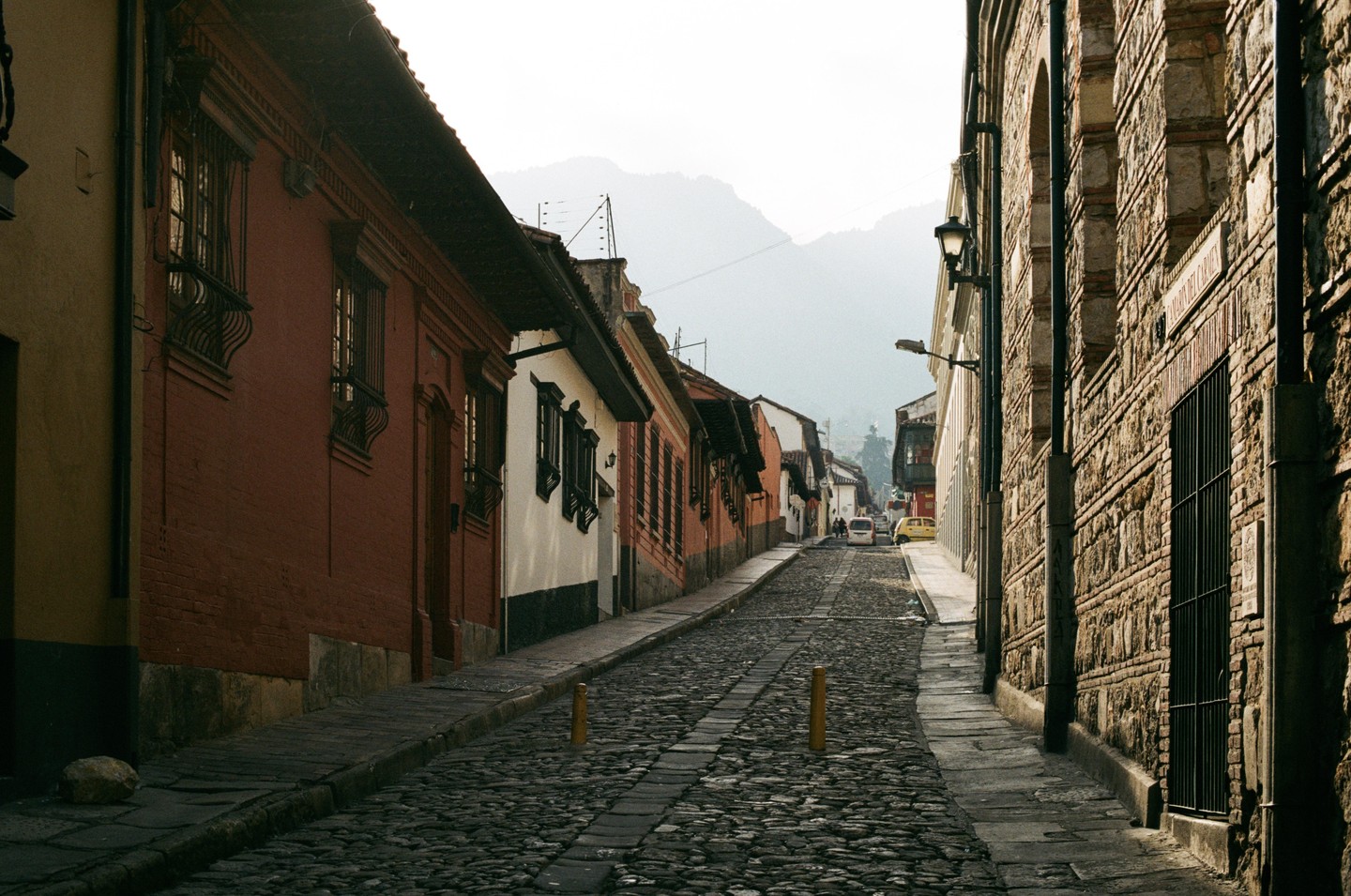 A narrow, cobblestone street lined with old buildings leads uphill towards distant mountains under a hazy sky.