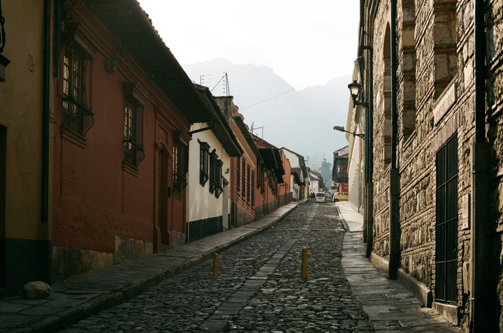 A narrow, cobblestone street lined with old buildings leads uphill towards distant mountains under a hazy sky.