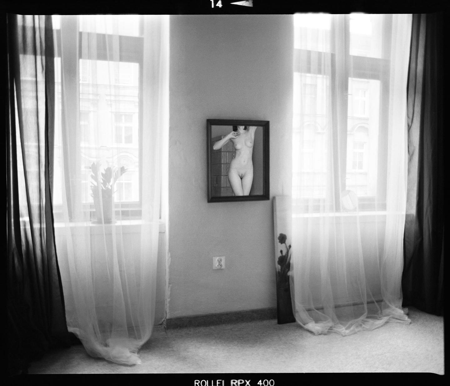A nude woman is reflected in a mirror on a wall between two windows with sheer curtains.