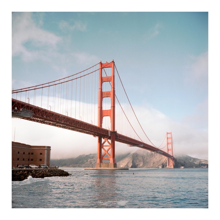 The Golden Gate Bridge spans across a body of water with a building and people on the shore.