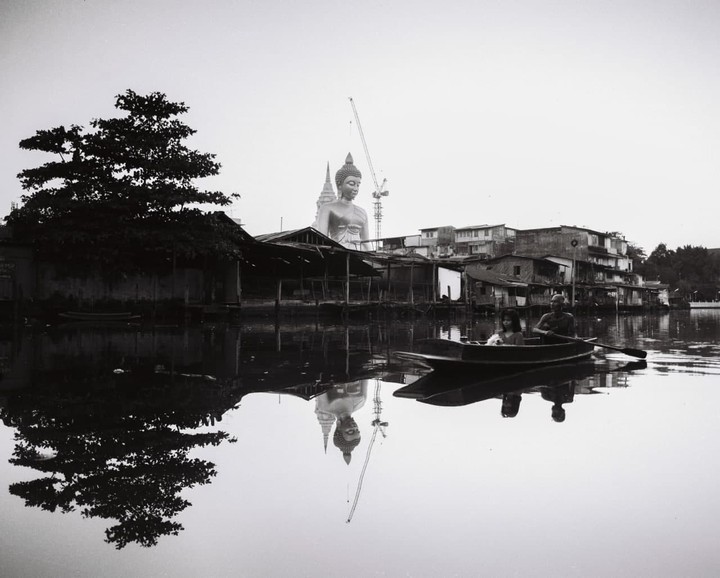 A man and a girl in a small boat row past a large Buddha statue and buildings on a river.