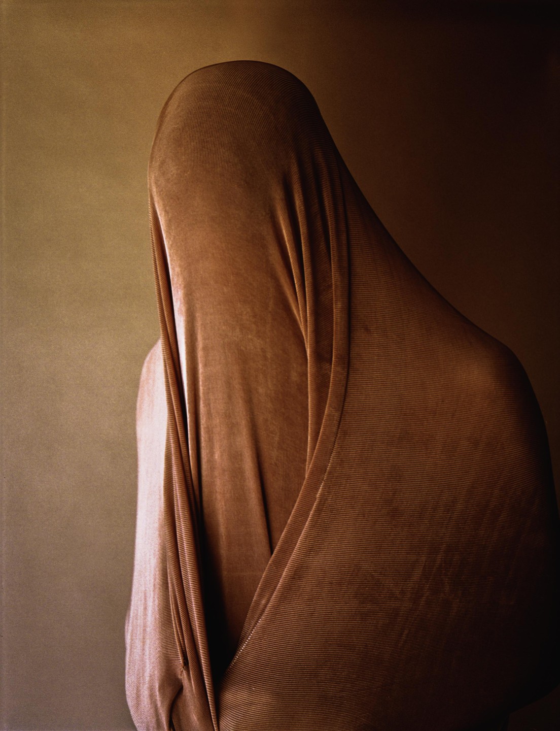 A figure is draped in a textured, brown fabric against a warm-toned background.