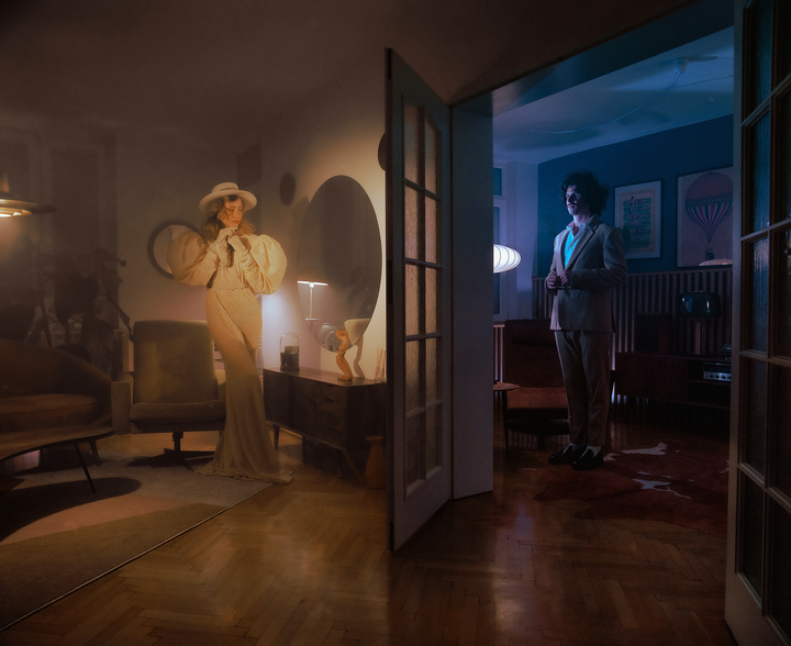 A woman in a white dress and hat stands in a dimly lit room while a man in a suit stands in a blue-lit room, separated by an open doorway.