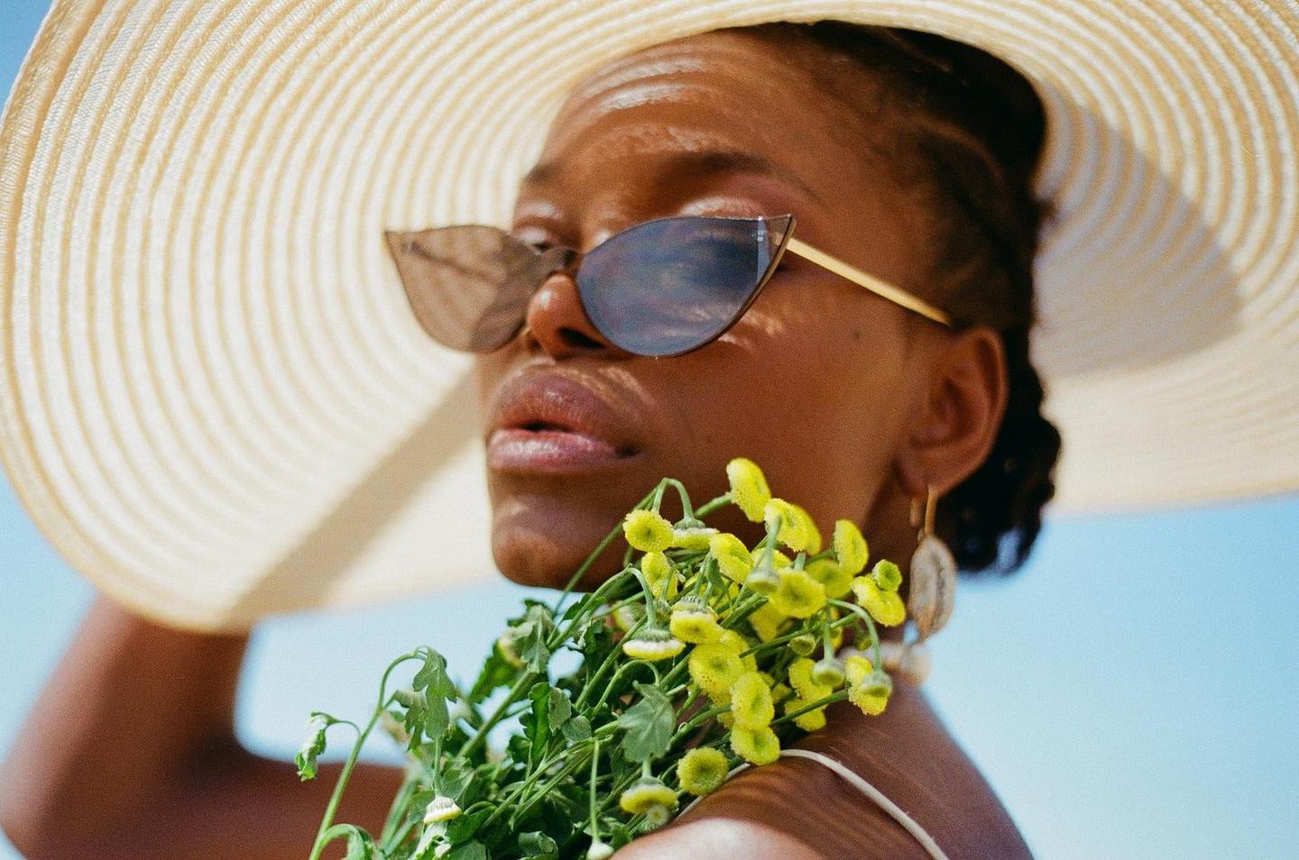 A woman wearing a wide-brimmed hat and sunglasses holds a bouquet of yellow flowers.