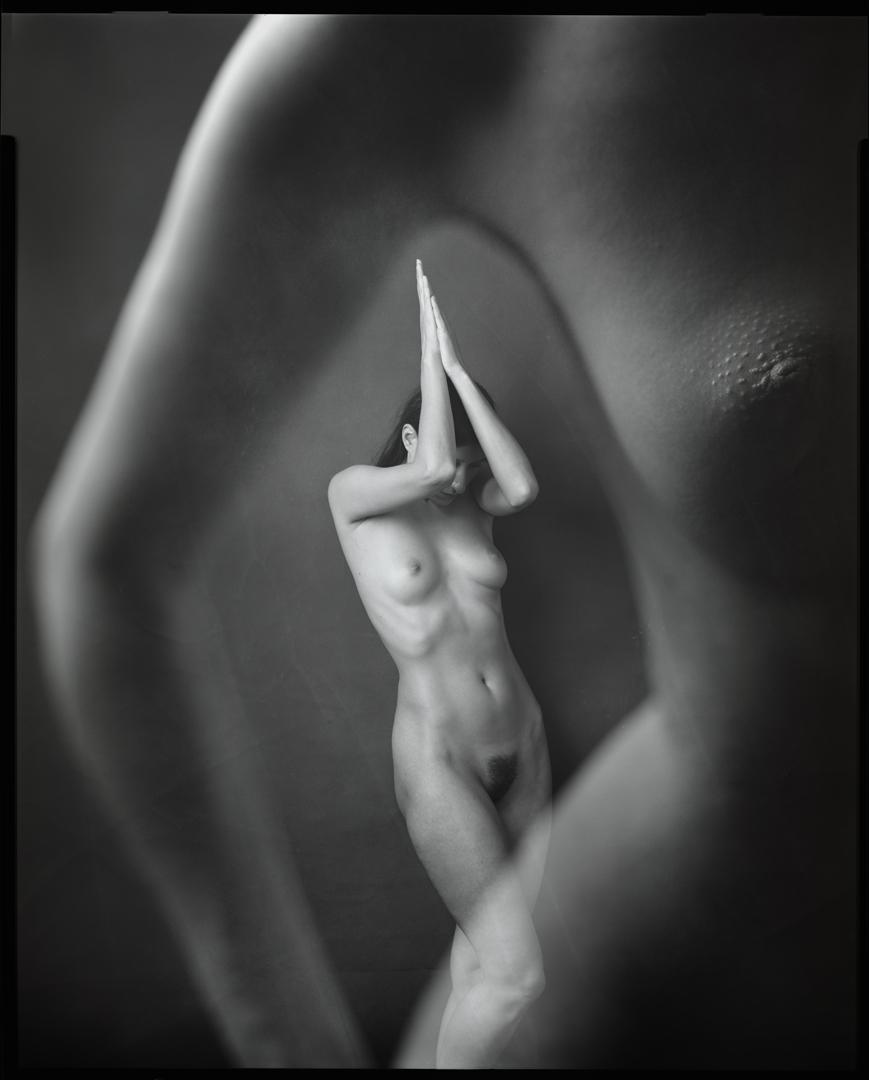 A nude woman stands with her hands together above her head, with a blurred nude body in the foreground.