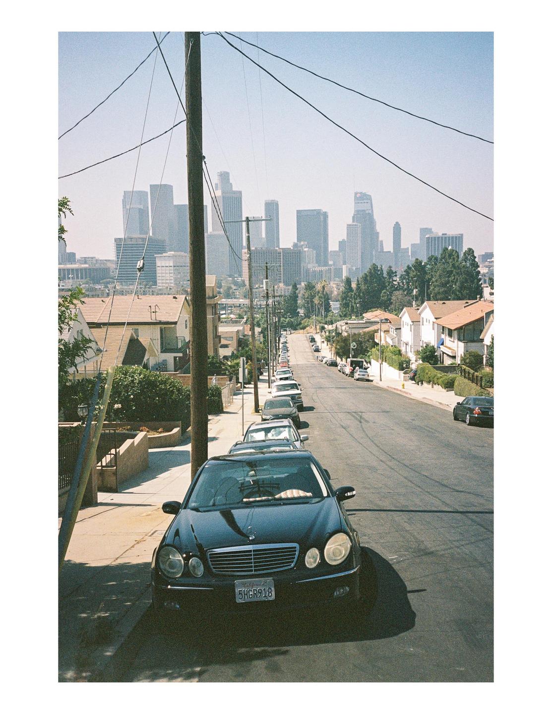A black car is parked on a residential street with a city skyline in the distance.