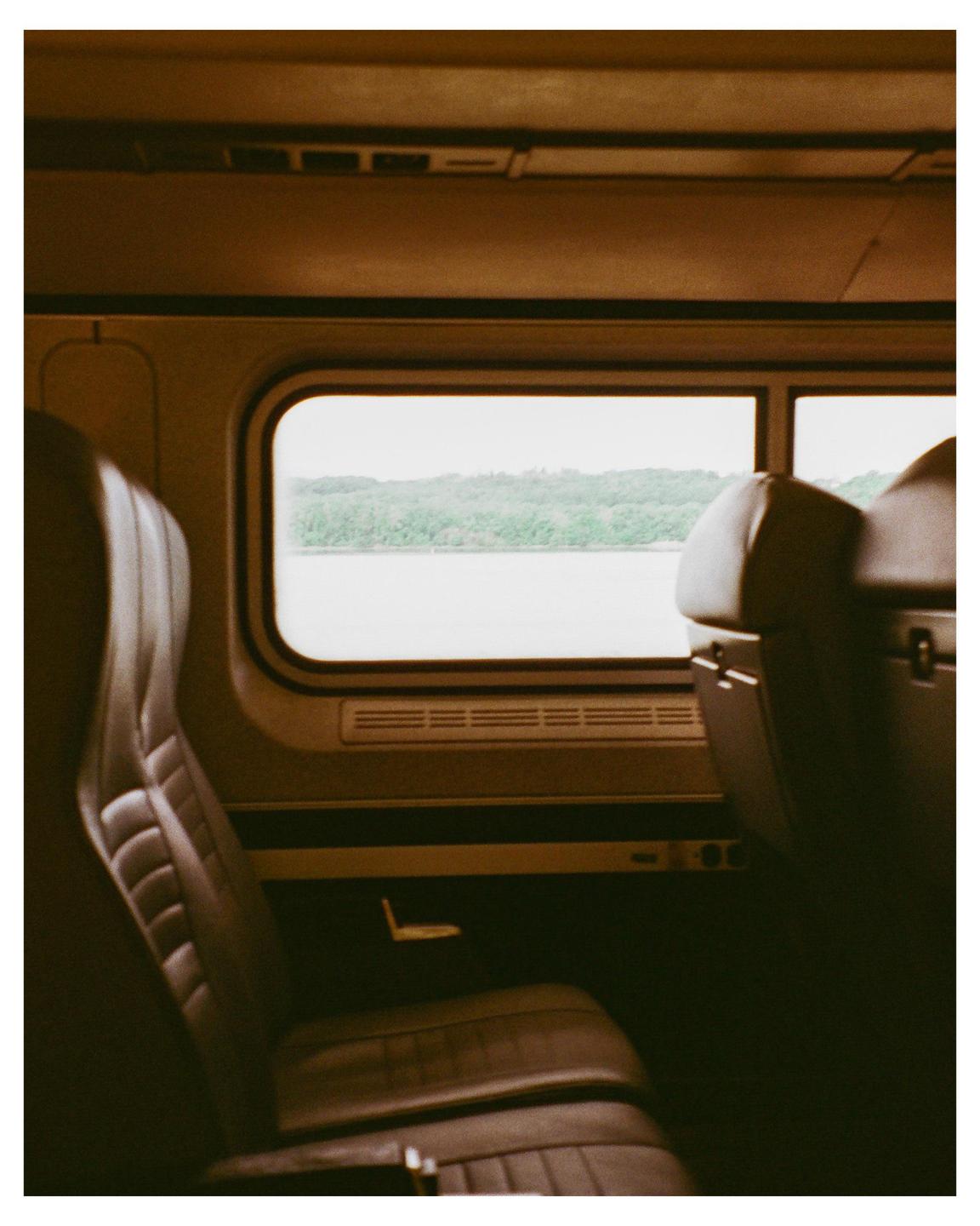 The interior of a train car shows two seats and a window view of a river and forested hills.
