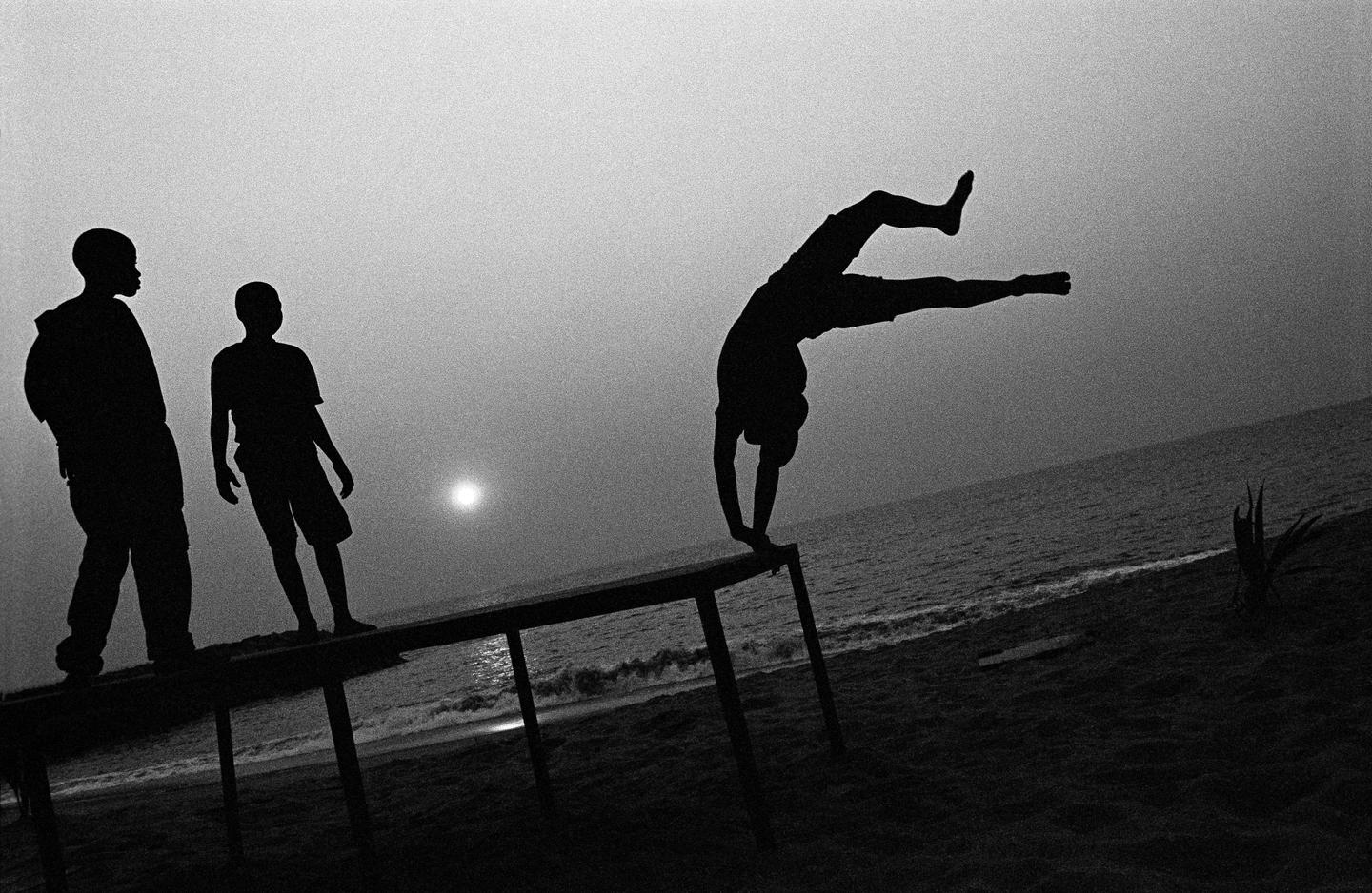 Three boys are silhouetted against the ocean and a bright sun, with one boy doing a handstand on a table.