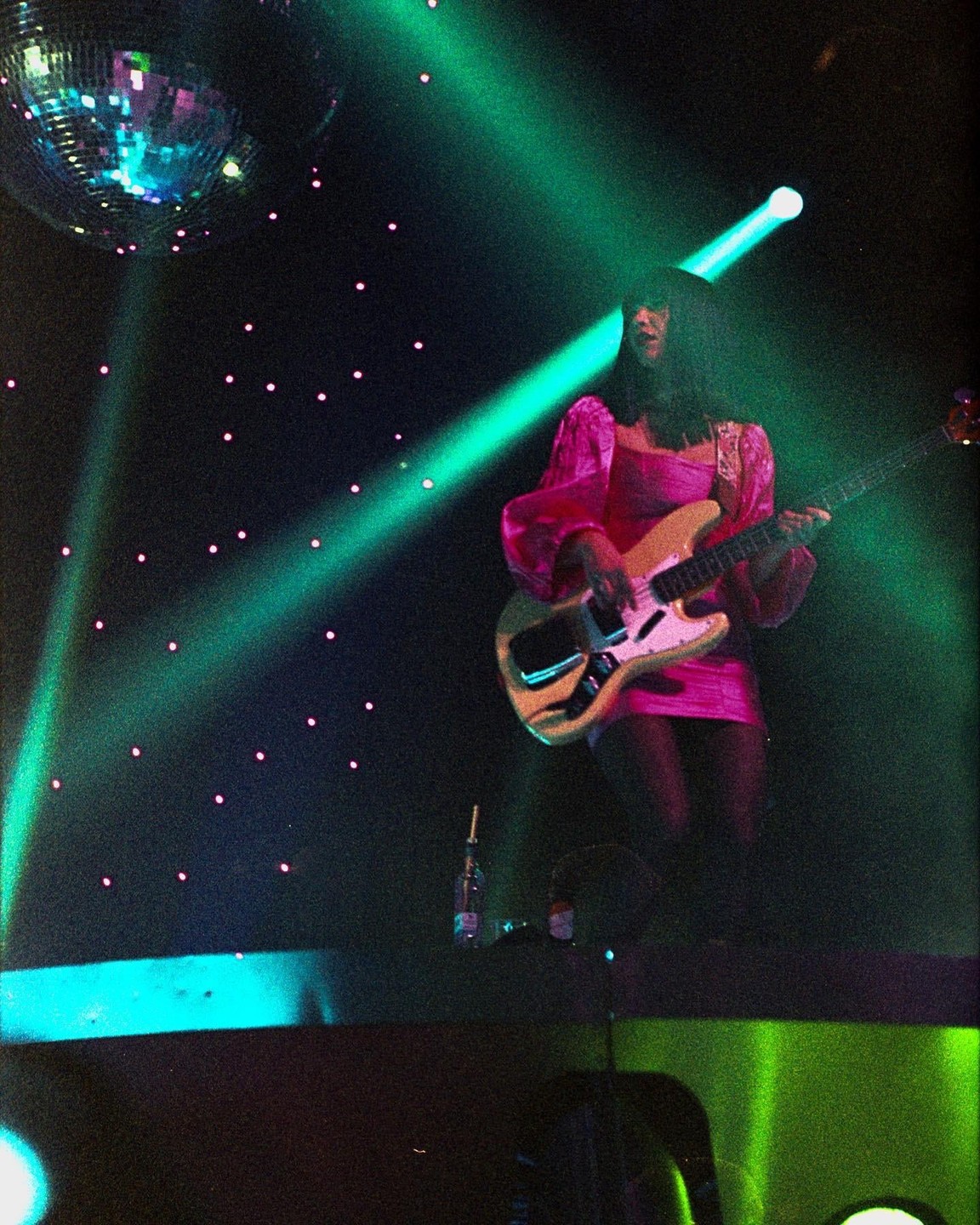 A woman in a pink dress plays a bass guitar on a stage with green lights and a disco ball.