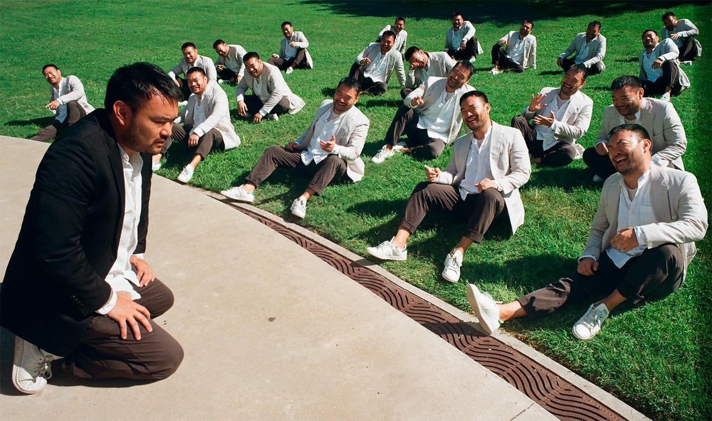 A man kneels on a concrete path while multiple versions of him sit on the grass behind him, laughing.