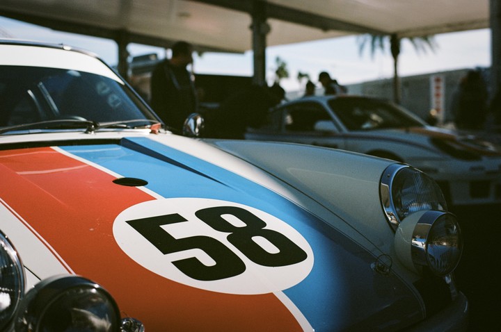 A vintage racing car with the number 58 on its hood is parked outdoors, with other cars and people visible in the background.