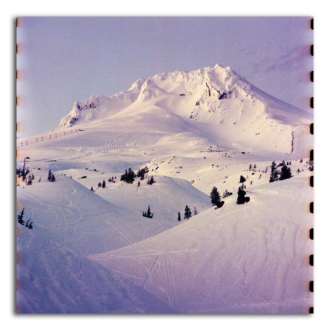 A snow-covered mountain with ski tracks and evergreen trees is visible under a clear sky.