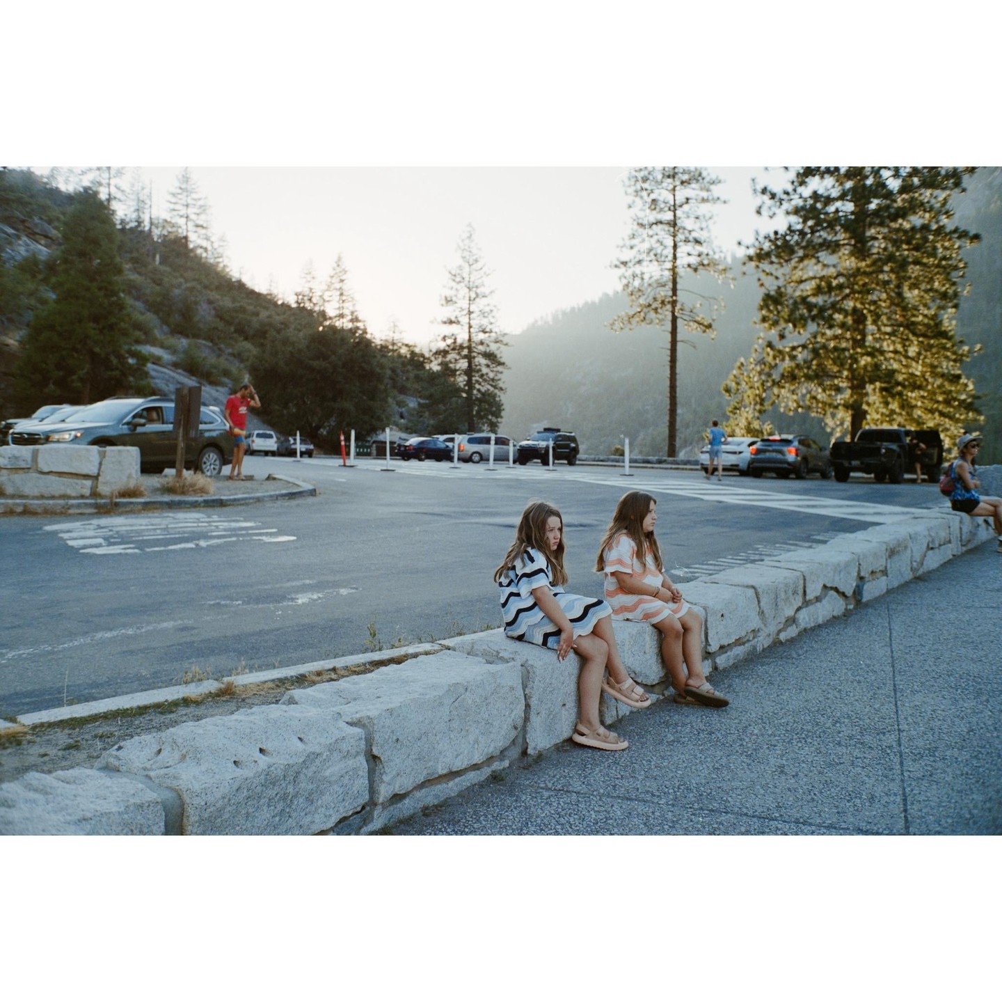 Two girls sit on a stone wall in a parking lot with mountains and trees in the background.