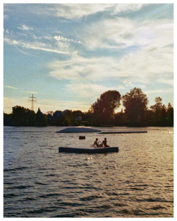 Two people sit on a floating platform in a lake with trees and a power line on the distant shore under a cloudy sky.